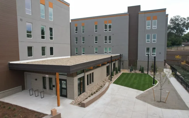 A modern, multi-story apartment building with gray and brown exterior, large windows, and a small landscaped courtyard with grass, sidewalks, young trees, and string lights.