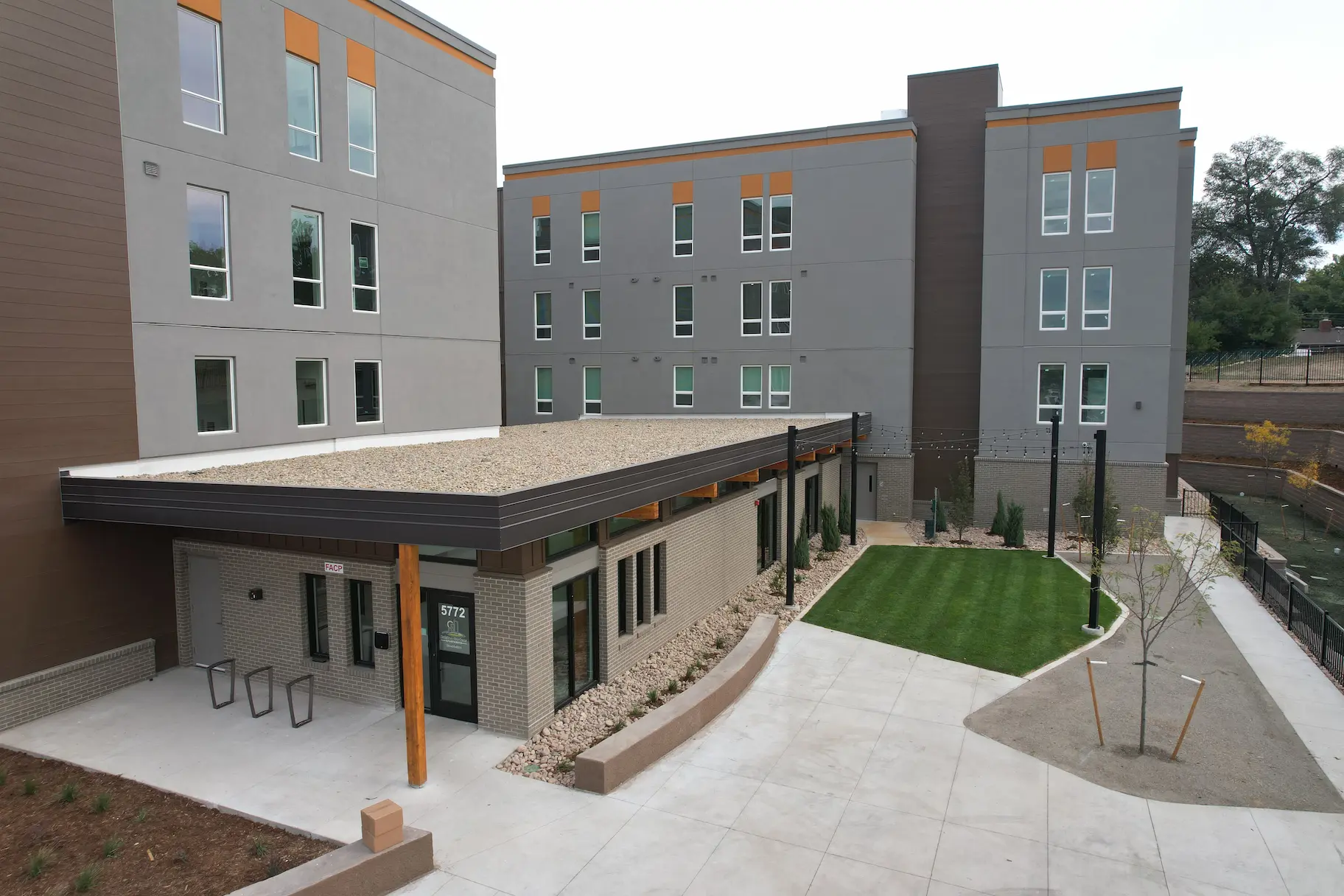 A modern, multi-story apartment building with gray and brown exterior, large windows, and a small landscaped courtyard with grass, sidewalks, young trees, and string lights.