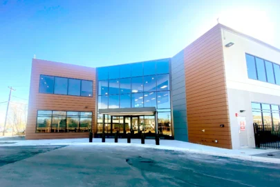 A modern two-story commercial building with large reflective glass windows and brown paneling, featuring a covered entrance and surrounded by a paved area under a clear blue sky.