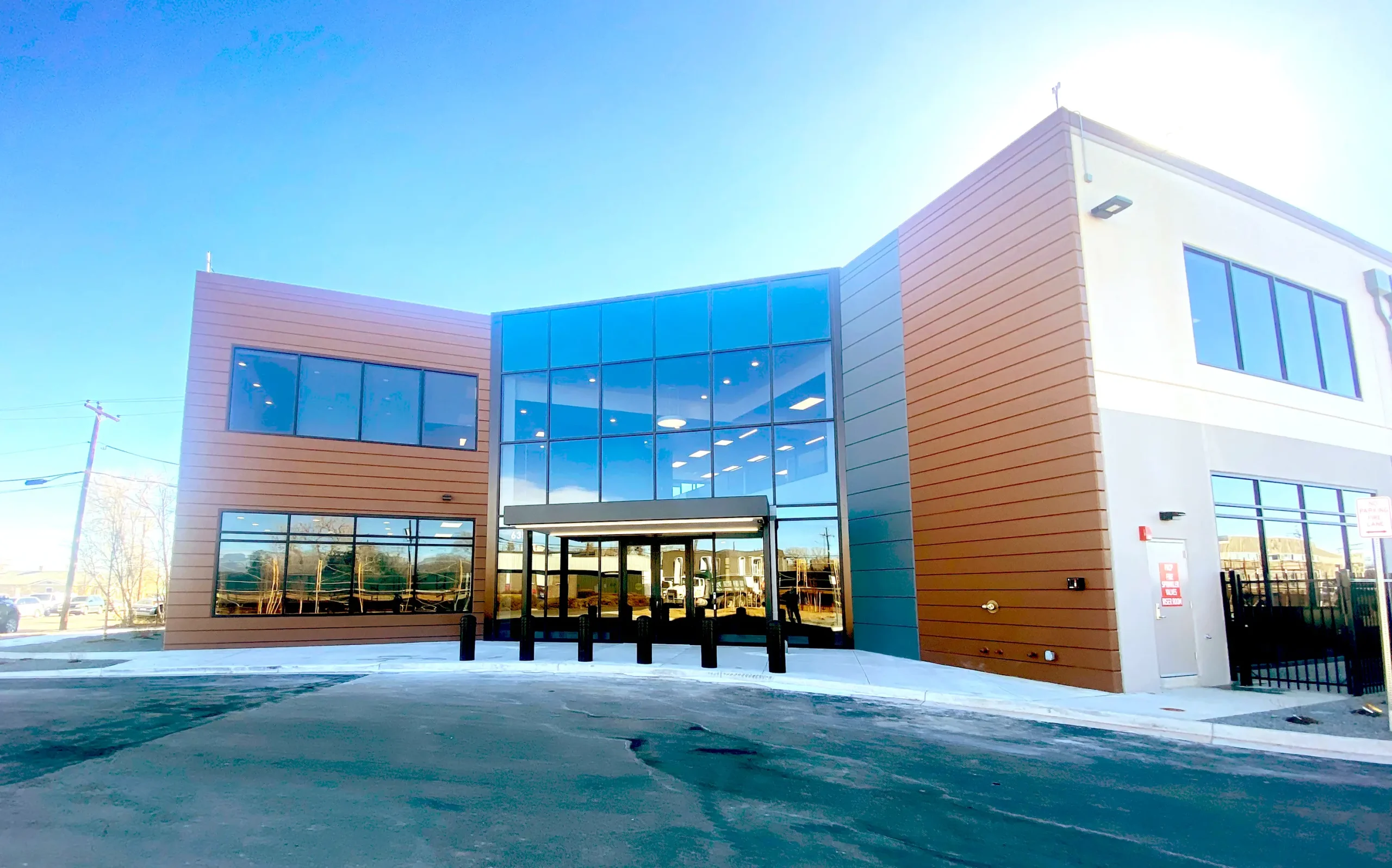 A modern two-story commercial building with large reflective glass windows and brown paneling, featuring a covered entrance and surrounded by a paved area under a clear blue sky.