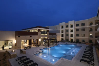 Outdoor hotel pool area at night, surrounded by lounge chairs and lit by soft lighting; modern multi-story hotel building in the background with large windows and balconies.