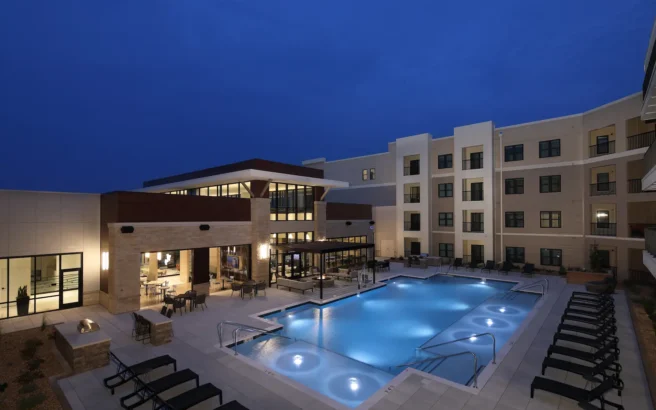 Outdoor hotel pool area at night, surrounded by lounge chairs and lit by soft lighting; modern multi-story hotel building in the background with large windows and balconies.