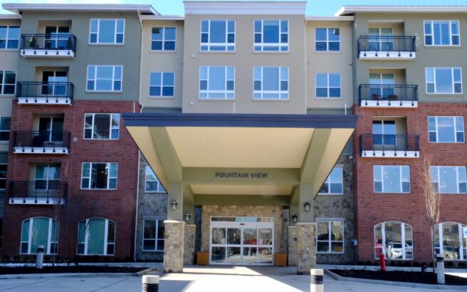 A modern, four-story apartment building with a large covered entrance labeled Fountain View. The facade features red brick and green siding with balconies and large windows.