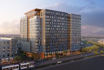 A modern high-rise apartment building with large windows and multiple floors stands by a busy street, surrounded by other buildings, trees, and parked cars under a partly cloudy sky.