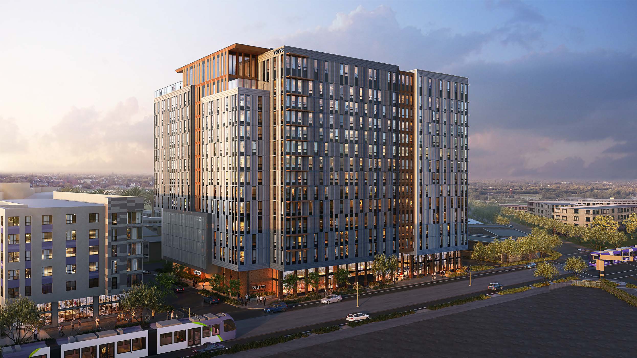 A modern high-rise apartment building with large windows and multiple floors stands by a busy street, surrounded by other buildings, trees, and parked cars under a partly cloudy sky.