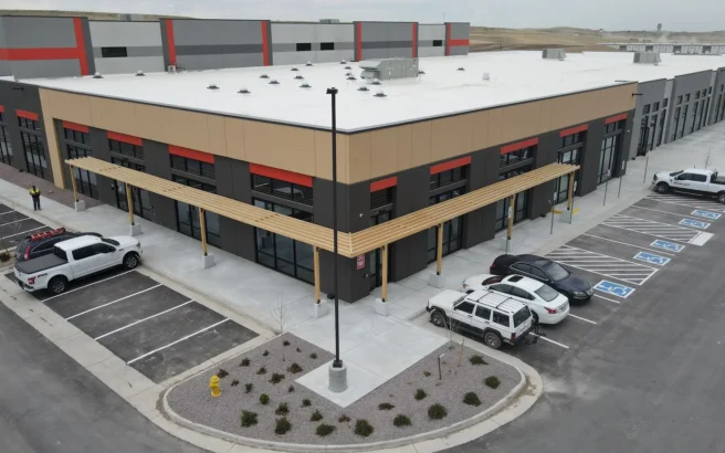 Aerial view of a modern commercial building with large windows, a beige and gray facade, and a white roof. Several cars are parked in front, including vehicles in accessible parking spaces. The building sits on a paved lot.