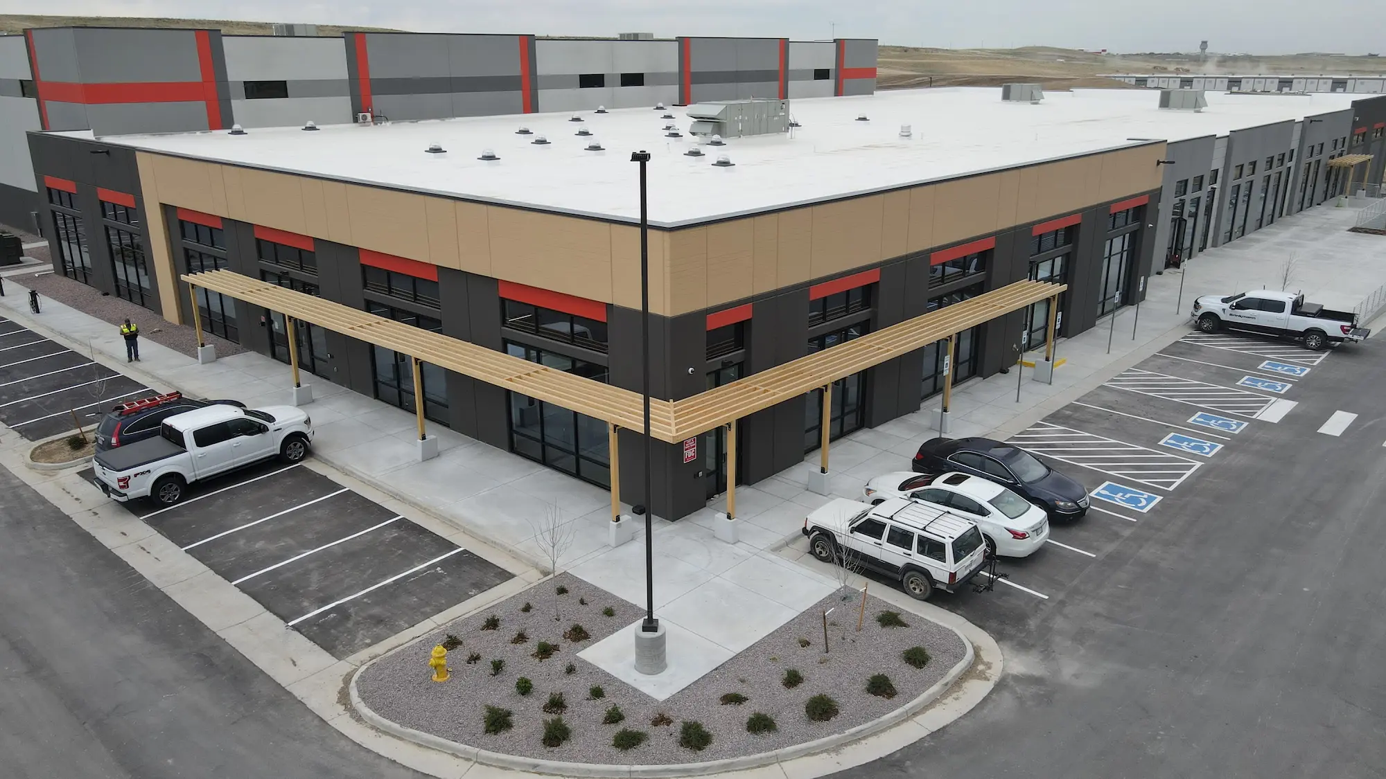 Aerial view of a modern commercial building with large windows, a beige and gray facade, and a white roof. Several cars are parked in front, including vehicles in accessible parking spaces. The building sits on a paved lot.