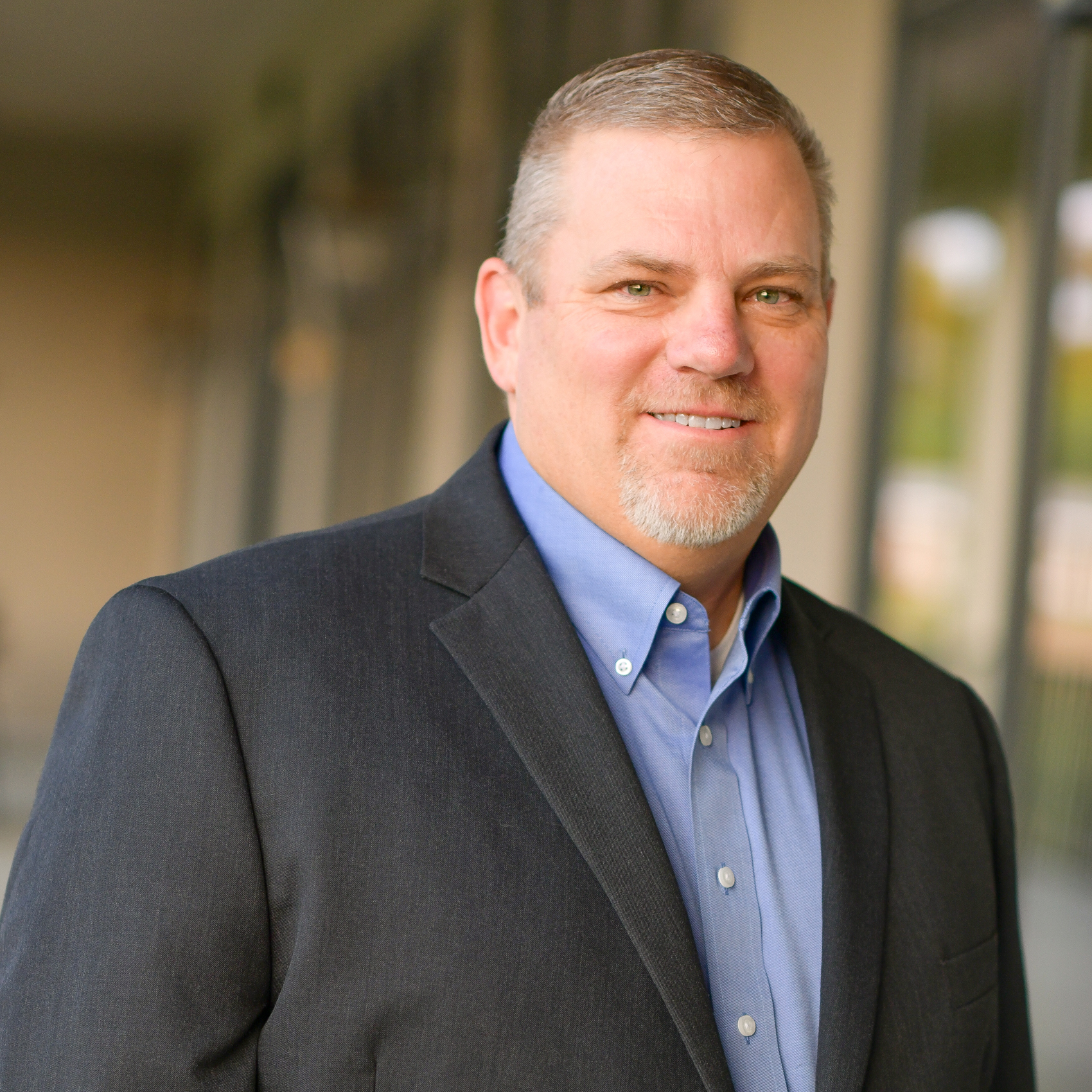 A middle-aged man with short light hair and a goatee is wearing a dark suit jacket over a blue dress shirt, smiling, and standing in a softly lit outdoor corridor.