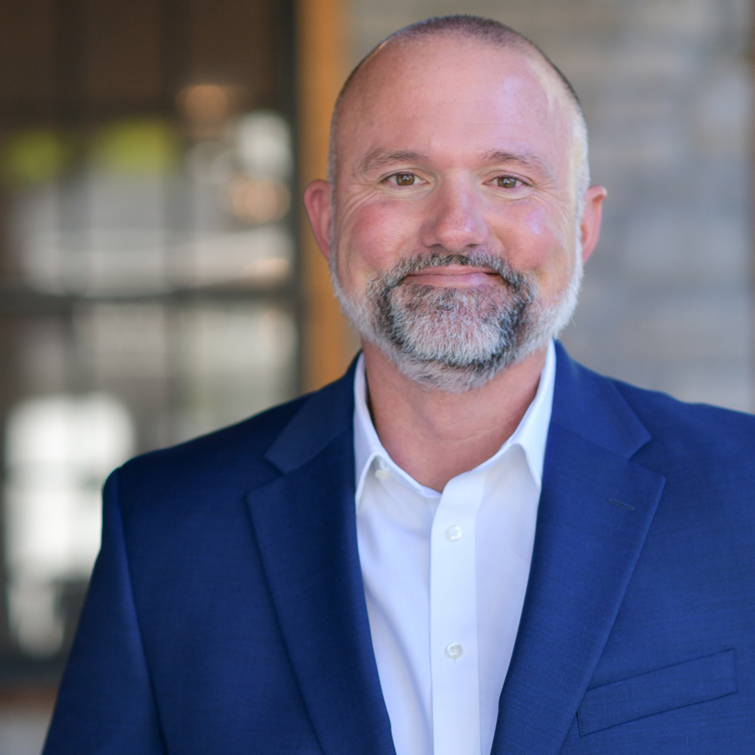 A middle-aged man with a trimmed gray beard and short hair smiles while wearing a blue suit jacket and white dress shirt, standing in front of a blurred background with windows.