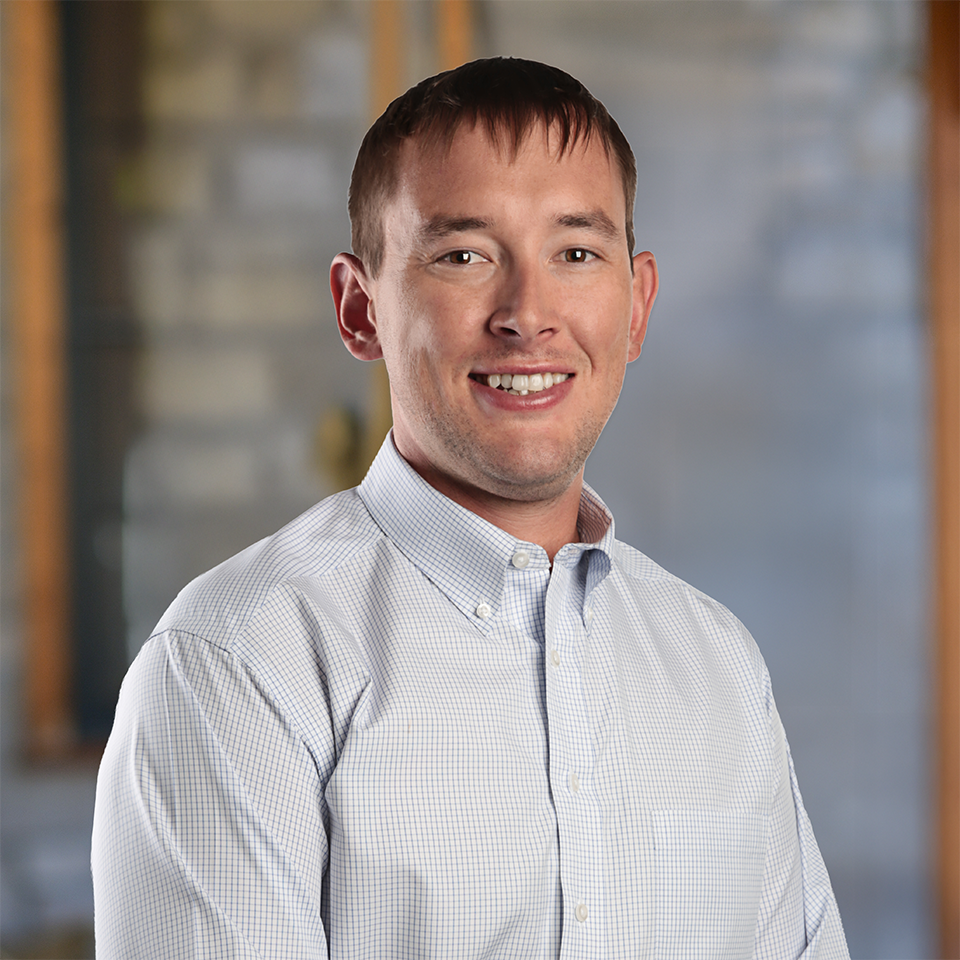 A young man with short brown hair smiles at the camera. He is wearing a light-colored, button-up shirt and stands in front of a blurred indoor background with gray and brown tones.