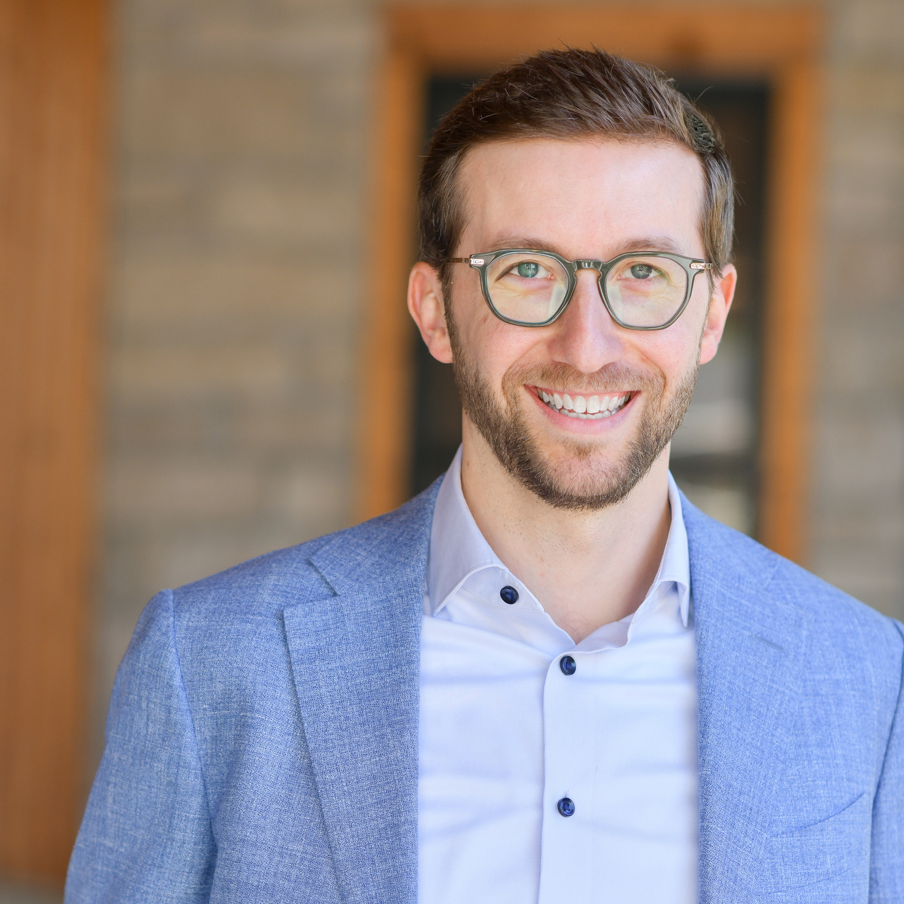 A smiling man with short brown hair, glasses, and a beard wears a light blue blazer and white shirt, standing in front of a blurred brick wall background.