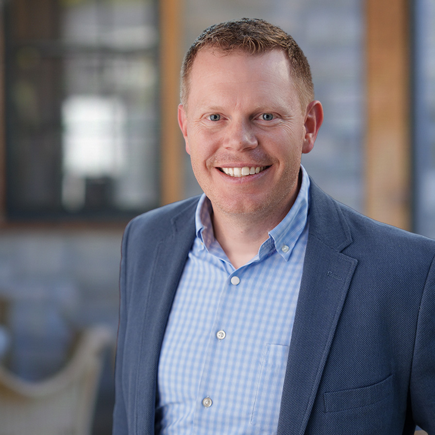 A smiling man in a blue checked shirt and blazer stands indoors with a blurred background featuring large windows and soft lighting.