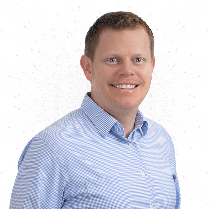 Smiling man with short light brown hair wearing a light blue checkered shirt, standing against a white background with a subtle pattern of connected dots.