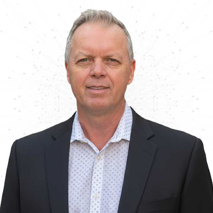 A middle-aged man with short gray hair wearing a dark suit jacket over a light-colored patterned shirt, standing against a white background with subtle dotted patterns.
