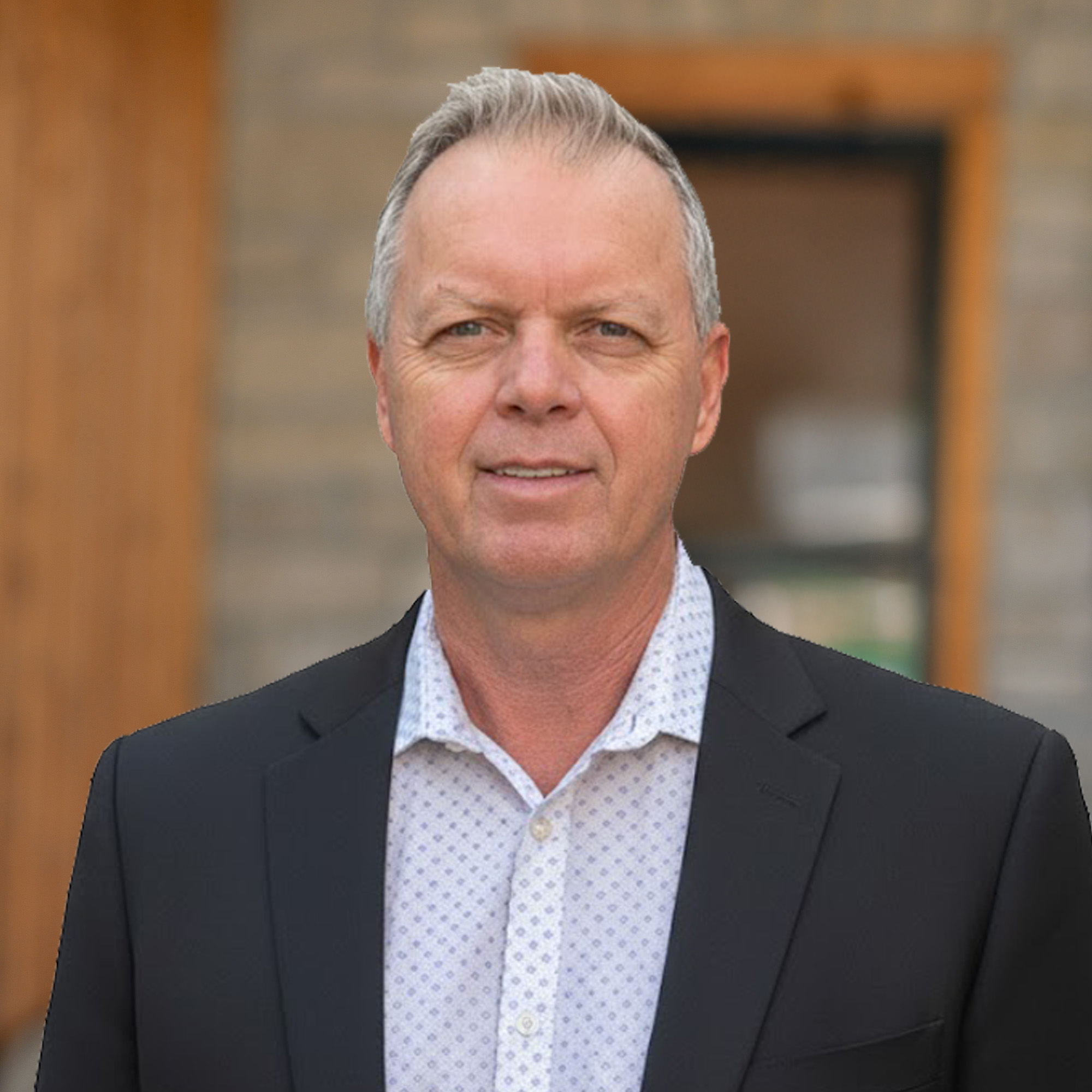 A middle-aged man with short gray hair wearing a black blazer and a white patterned shirt stands outside in front of a blurred brick and wooden background.