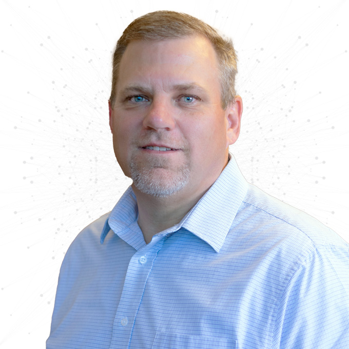 A middle-aged man with short light brown hair and a goatee, wearing a light blue collared shirt, stands in front of a white background with subtle abstract patterns.